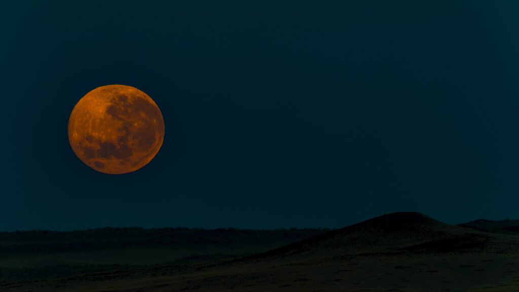 Full moon over a hills. It's orange and deep in colour. The background is blue. 