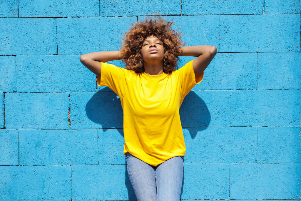 Black woman with a gorgeous yellow t-shirt and jeans leaning against a stark cerulean blue brick wall.
