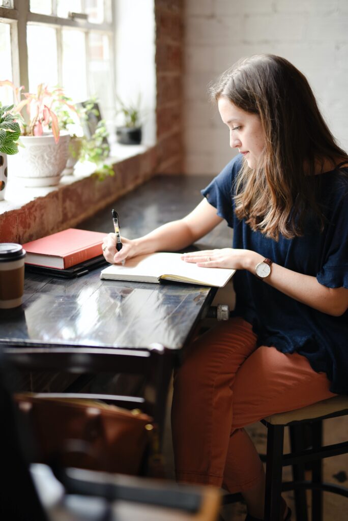 Brunette woman sitting on a bar in a cafe near a plant-lined window writing in a journal. A coffee cup and a stack of books sits next to her. 