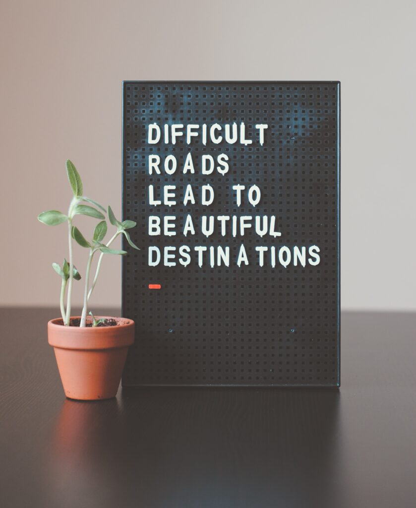 Peg board with message on it sitting beside a cute little plant. The message says "Difficult Roads Lead to Beautiful Destinations."